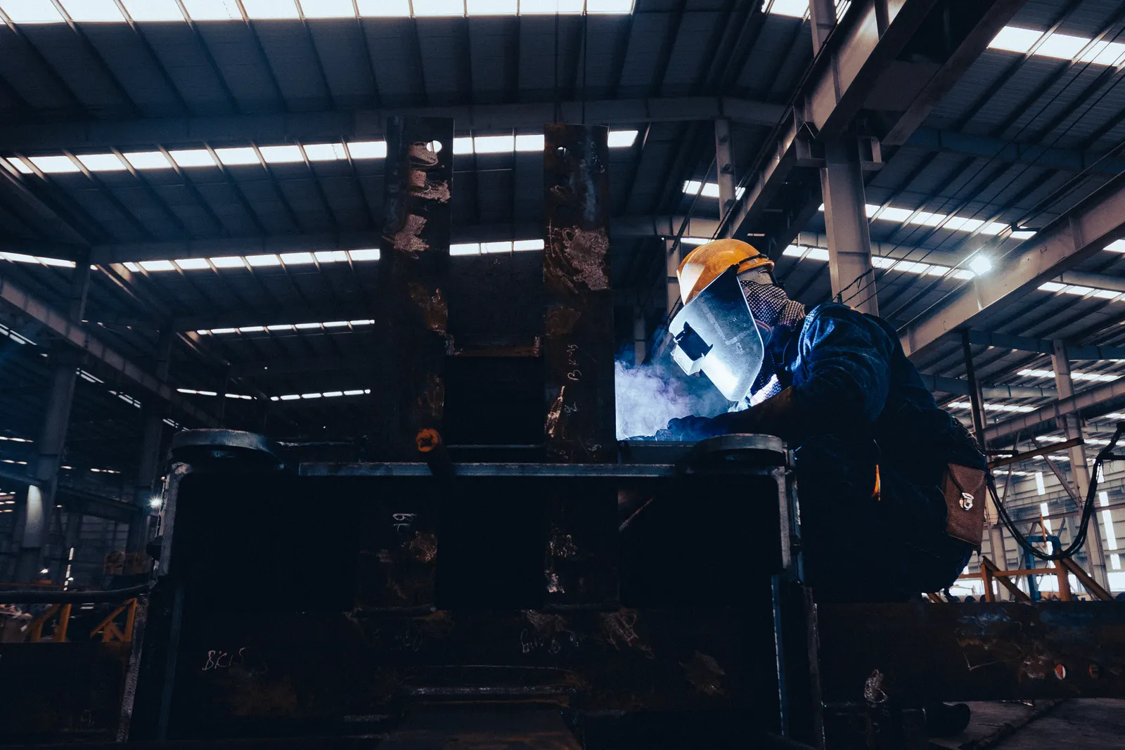 Welder in a hard hat and face shield welding a steel assembly in a fabrication shop